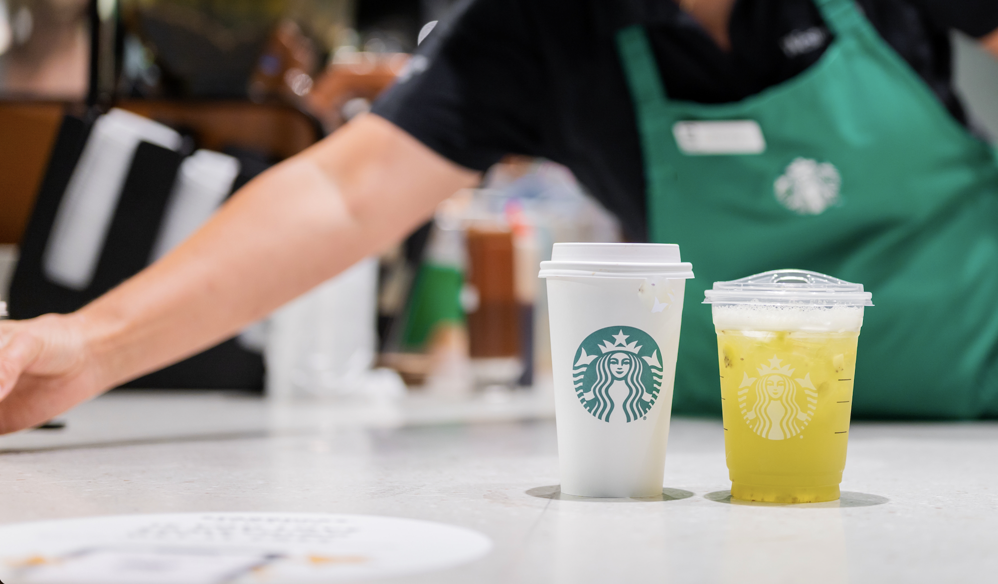 A barista standing behind a hot coffee and lemonade refresher on the counter.