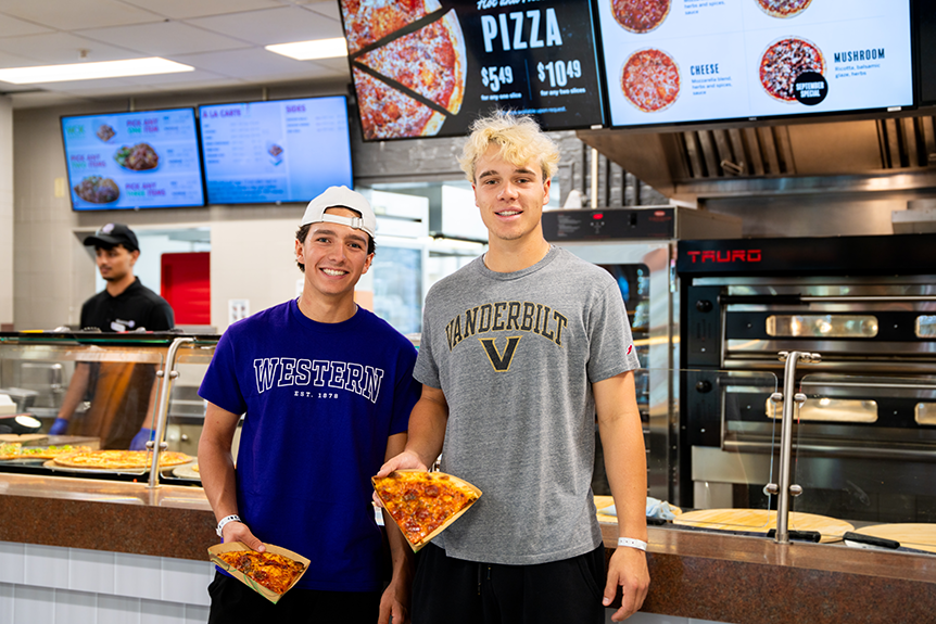 Two students holding slices of pizza in front of the Pizza eatery in UCC Food Hub.